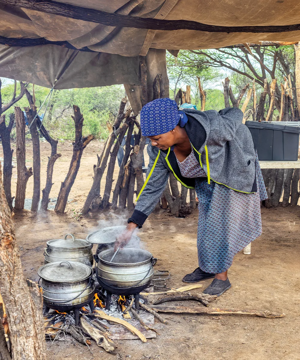 A woman cooks over an open wood fire in a dark, smoke-filled kitchen.