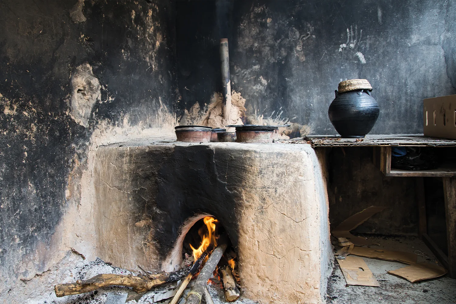 A woman cooks over an open wood fire in a dark, smoke-filled kitchen.