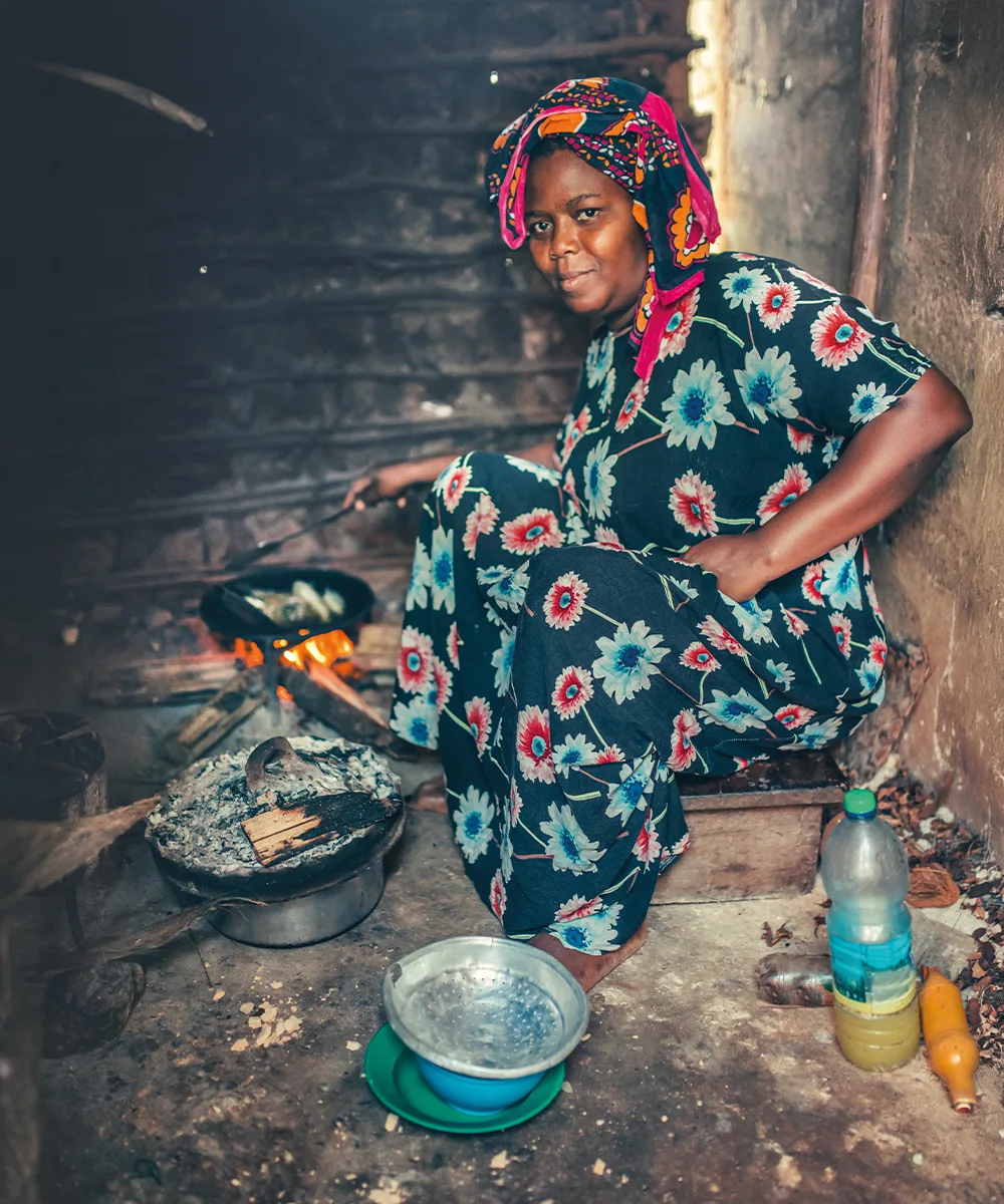 A woman cooks over an open wood fire in a dark, smoke-filled kitchen.